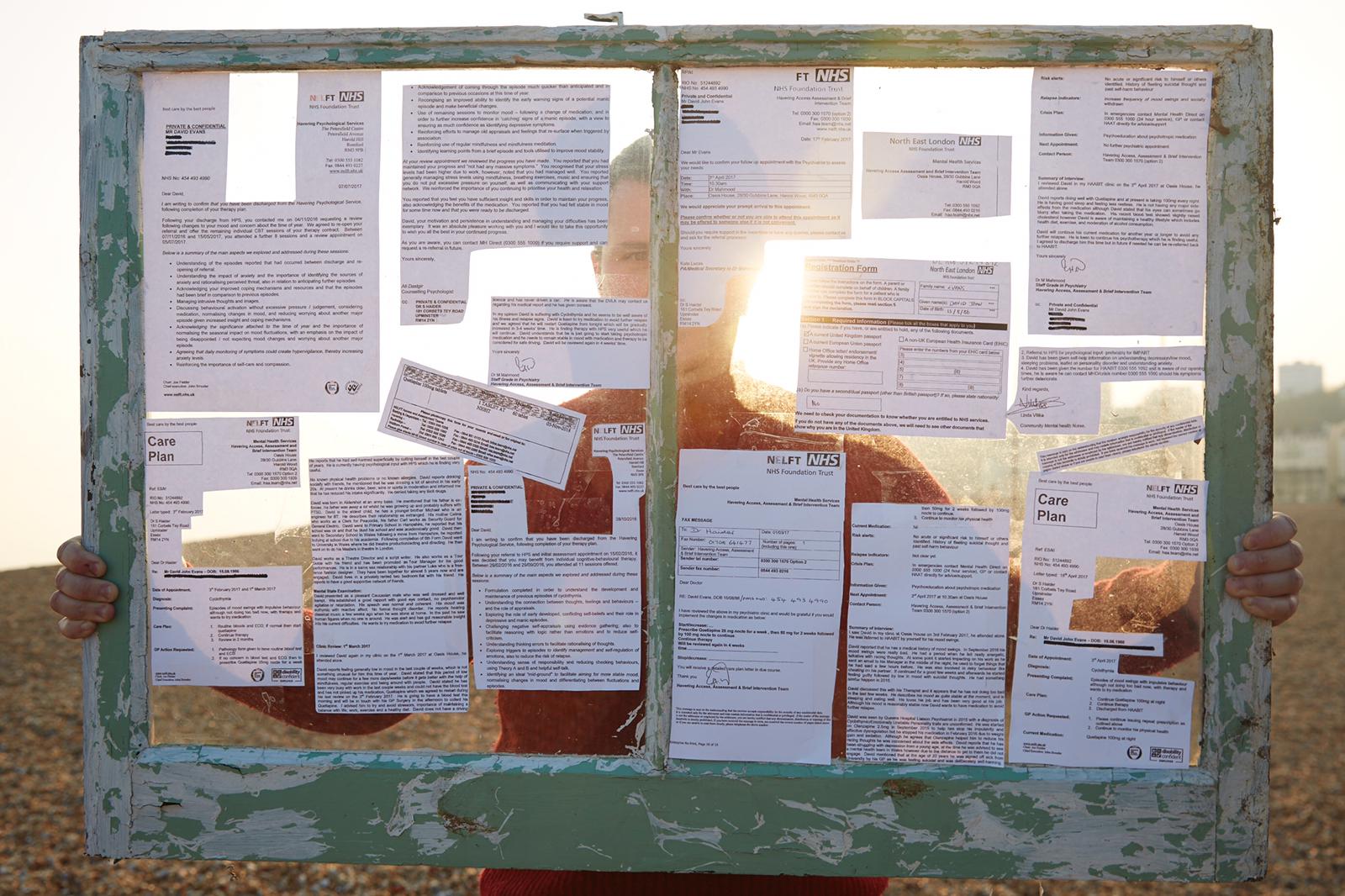 dewi evans, standing on a beach holding a window which is pasted with letters from his psychiatrist and councilors